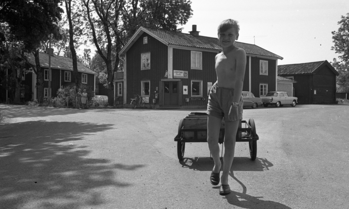 Lekhyttan 21 juni 1966

En pojke drar en lastkärra efter sig i Lekhyttan. Han är klädd i kortbyxor och tofflor. I bakgrunden syns bl.a. Lekhyttans handel, andra byggnader, två äldre damer samt två bilar.