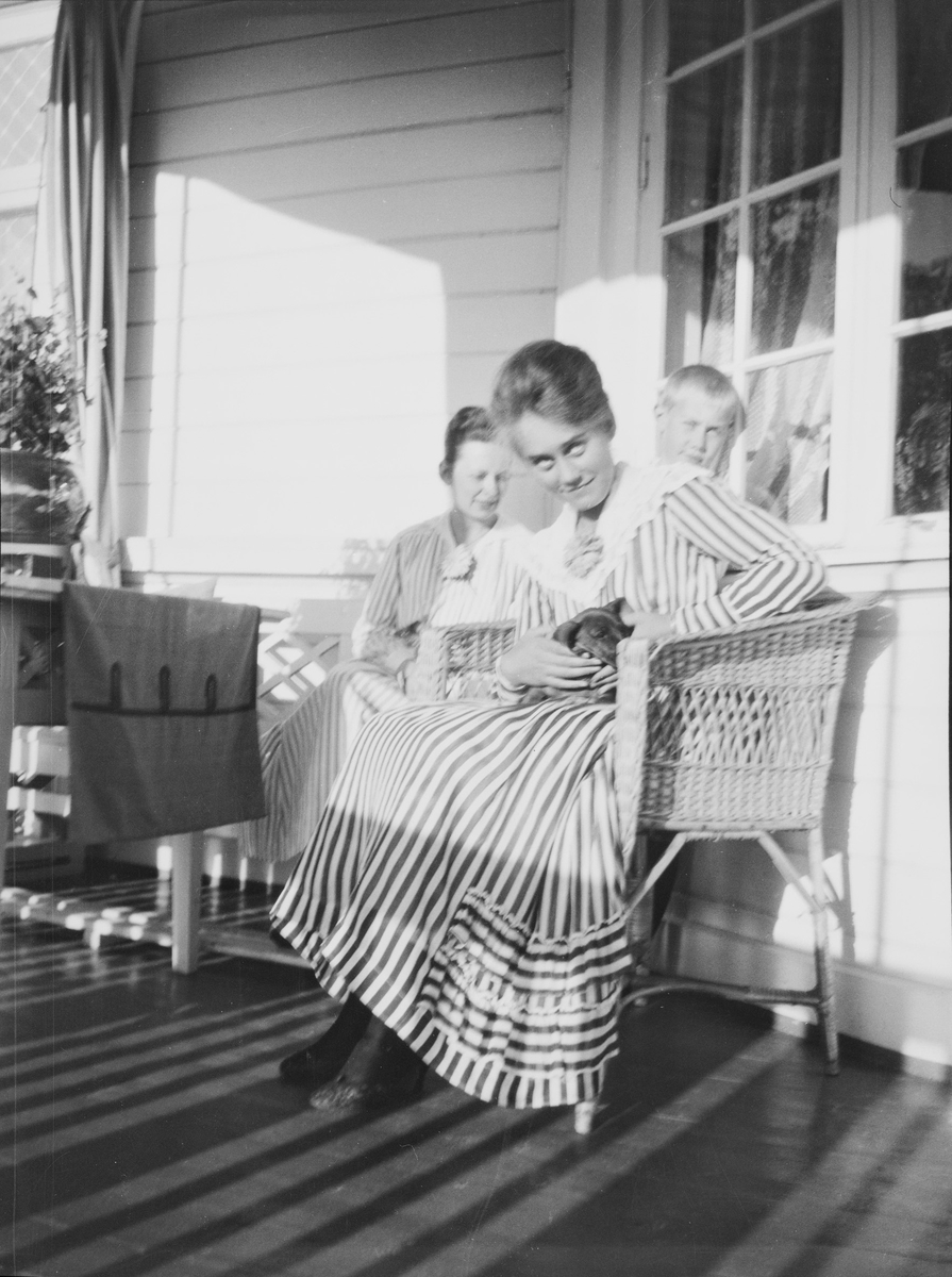 Fra venstre: en uidentifisert kvinne, Ise (Louise) Mathiesen med en liten dachs på fanget og broren Iacob bak, sitter på veranden.