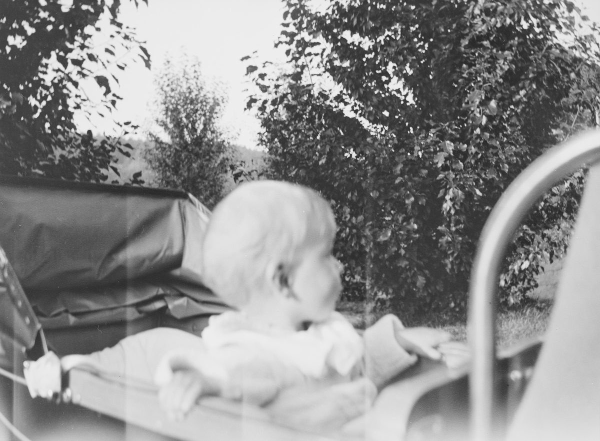 Et lite barn, lille Pierre, sitter i en barnevogn som står på gårdsplassen til Linderud Gård ved sommerstid. Barnet har vendt seg bort fra fotografen og ser ut til å følge med på noe til siden for ham.