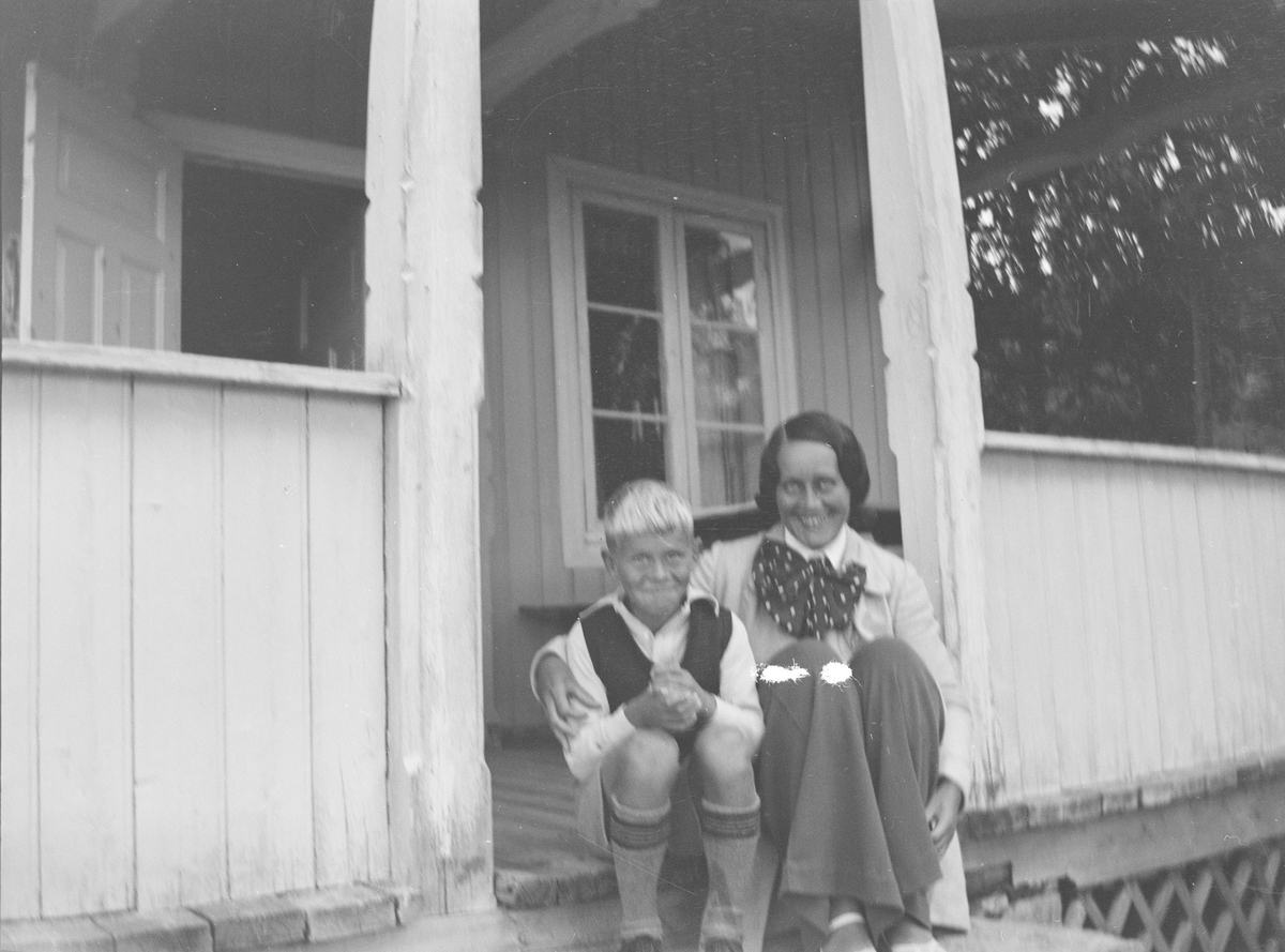 En gutt og en kvinne, Ole og Jil, sitter på trappen opp til verandaen til et hus i Jeppedalen. Begge smiler til fotografen. Døren fra verandaen inn til huset står åpen, det er sommer.