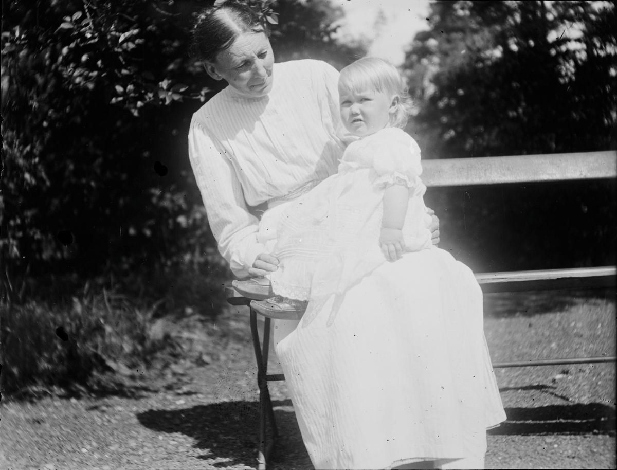 En eldre kvinne sitter på en benk i en hage med Iacob Ihlen Mathiesen på fanget.