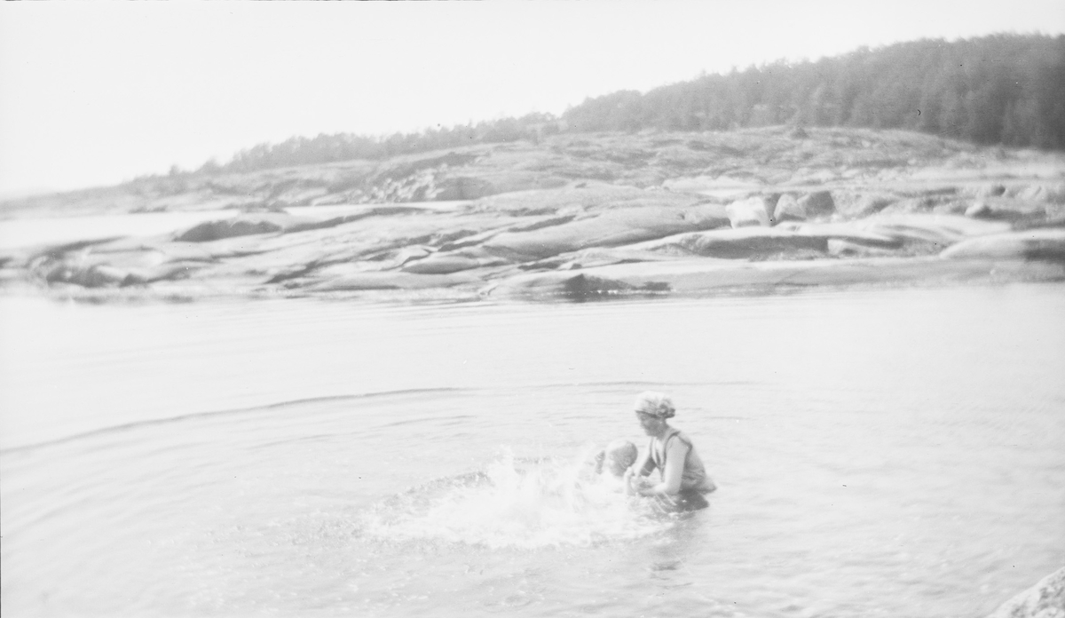 En kvinne står i vannet og holder en plaskende gutt under armene, som hjelp til å lære å svømme på rygg. De står i vannet nesten til livet. Bak kvinnen og gutten ligger områder med svaberg før skogen tar over lenger inn på land.