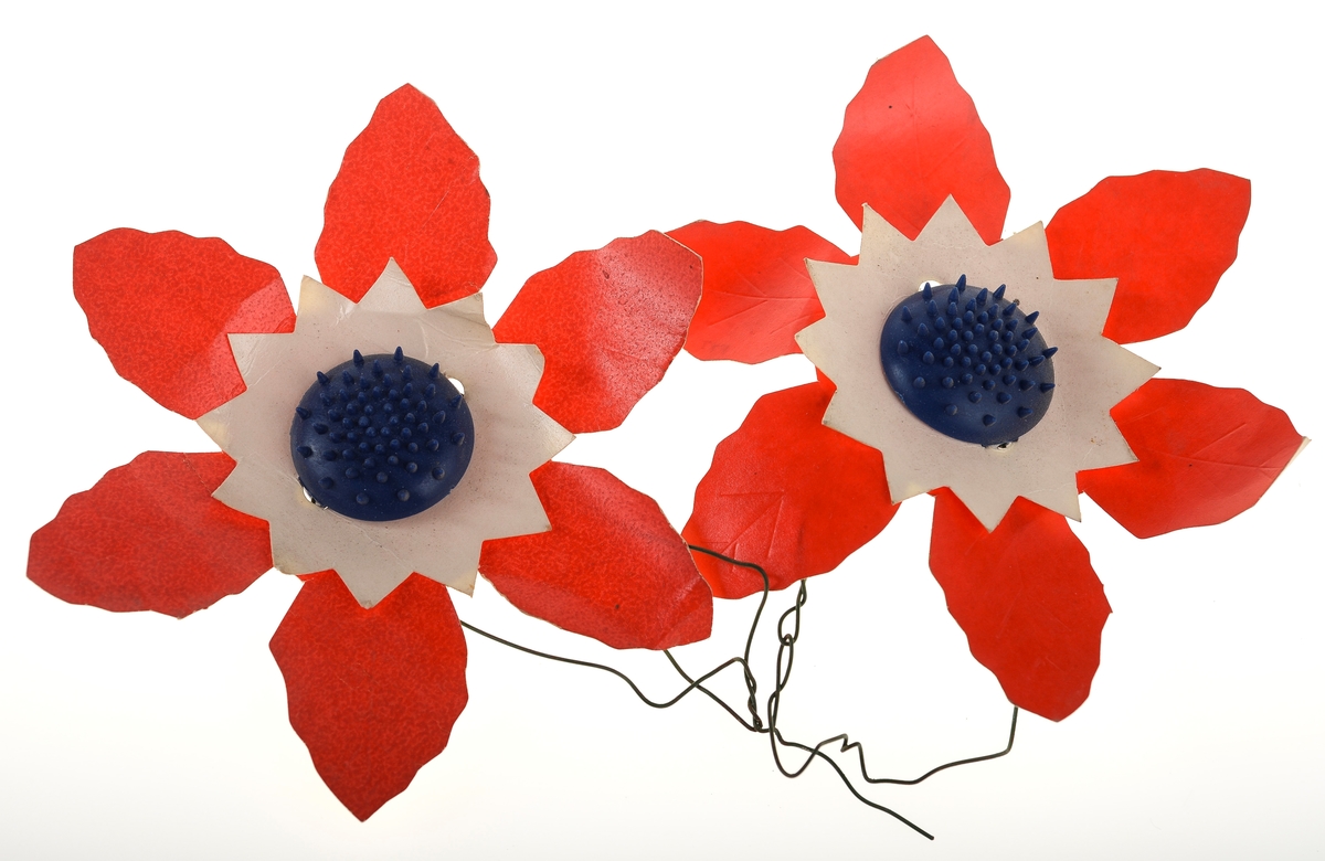 To identiske maiblomster. Blomsten består av to lag papp. Det nederste laget er seks røde kronblader, mens det øverste laget er hvit papp med stjerneform. Disse holdes sammen av en blå plastknapp. Grønn ståltråd er festet på baksiden.