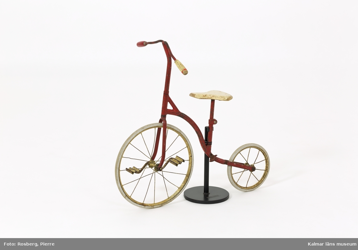 KLM 39651. Cykel, barncykel. Cykeln har två hjul, ett större fram och ett mindre bak. Tramporna sitter direkt på framhjulet. Stomme av rödmålat järn. Gummiklädda hjul. Sadel och handtag av trä, vitmålade. Ena handtaget är utbytt, rödmålat. Cykeln är monterad på ett stativ av järn.