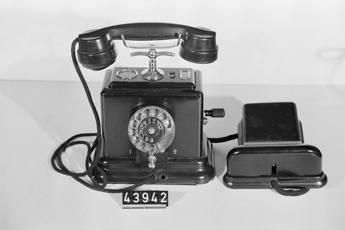 Telefonapparat för AT-system, mellanstationsapparat, 2 dl, med en anknytning. Bordsmodell med svartlackerad plåtkåpa, mikrotelefon av bakelit modell m33, fingerskiva av förnicklad mässing, 3-vägs omkastare, återfjädrande tryckomkastare, blänkare, apparatklocka, induktor med vev och väggplint med ringklocka. Den återfjädrande tryckknappen på apparatens ovansida har en dubbel funktion - vid signalgivning med vevinduktorn tystas den egna apparatklockan och under ett samtal kan mikrofonen temporärt kortslutas för att förbättra hörbarheten i hörtelefonen vid lyssning.