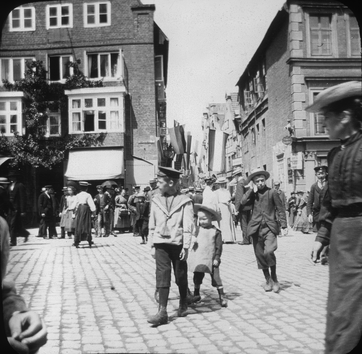 Skioptikonbild med motiv av tre små pojkar i folkvimmel på torg i Lüneburg.
Bilden har förvarats i kartong märkt: Resan 1906. Lüneburg 8.