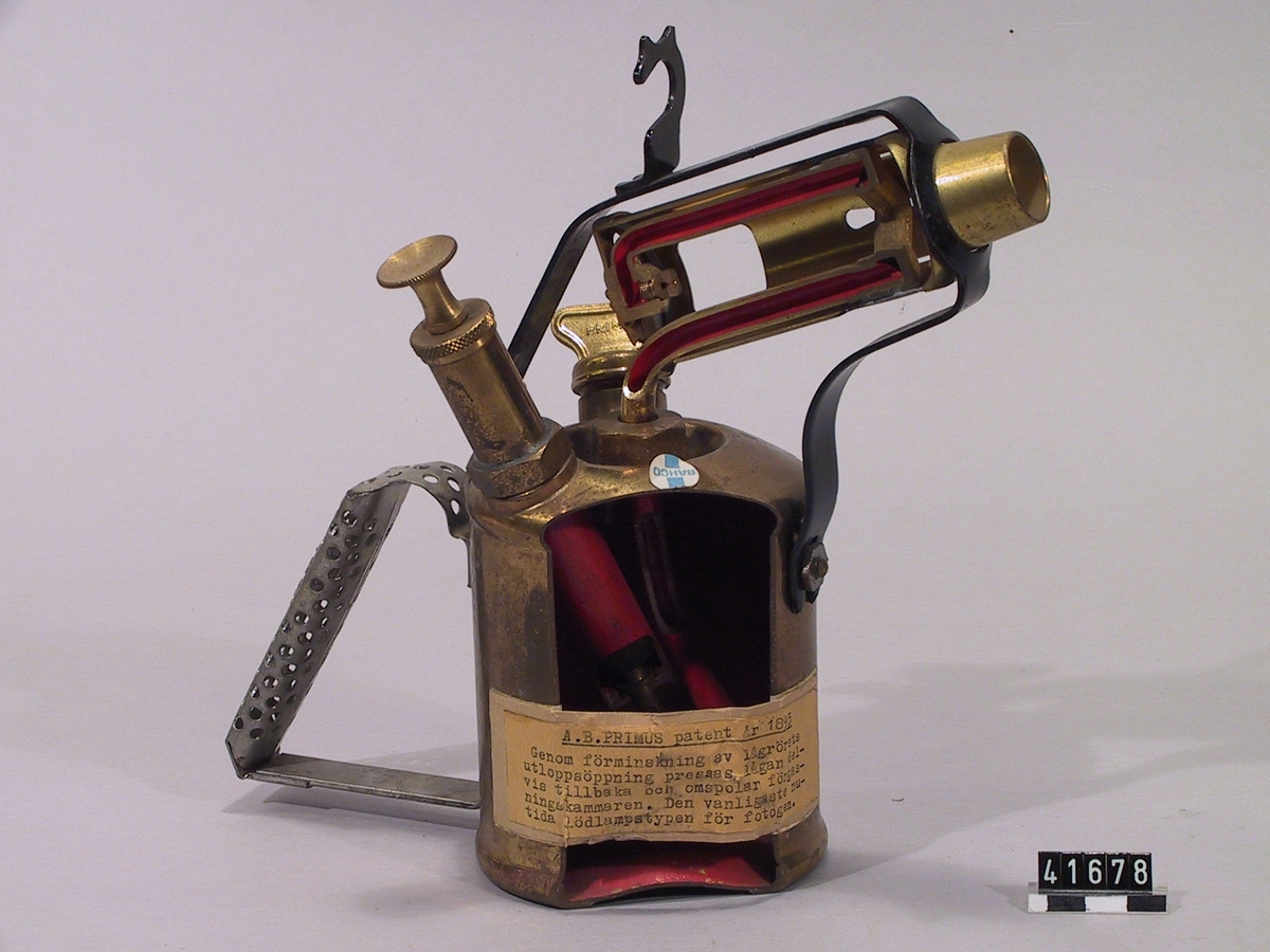 Lödlampa av mässing, itusågad för utställning. Genom förminskning av lågrörets utloppsöppning pressas lågan delvis tillbaka och omspolar förgasningskammaren. Den vanligaste nutida lödlampstypen för fotogen. Etikett med text: AB Primus patent år 1893.
