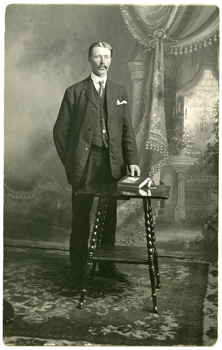 Portrett av mann i helfigur stående ved et bord. Han er kledd i dress og har slips og lommeur. Bildet er tatt i et atelier der lerretet viser et interiør. Kan det være fra Amerika?
