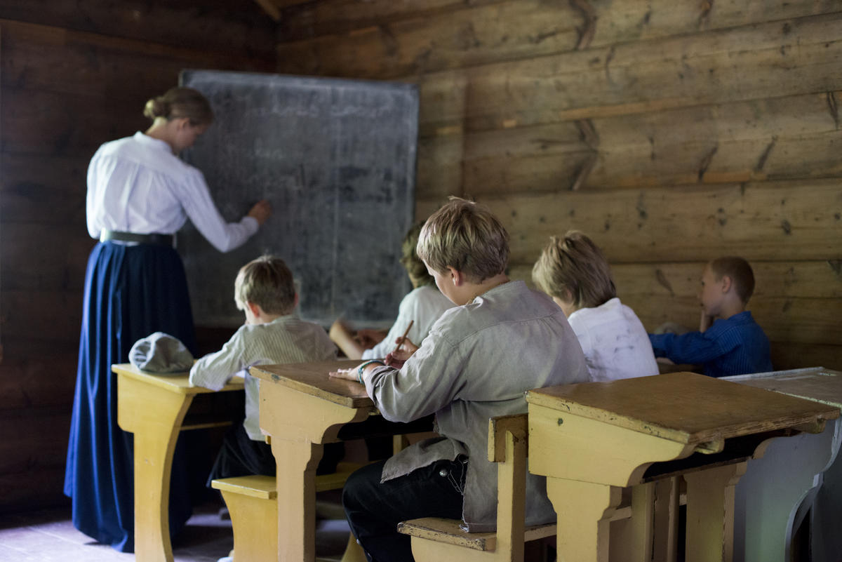 Barn sitter ved pultene i gammel skolstue mens lærerinne skriver på tavlen
