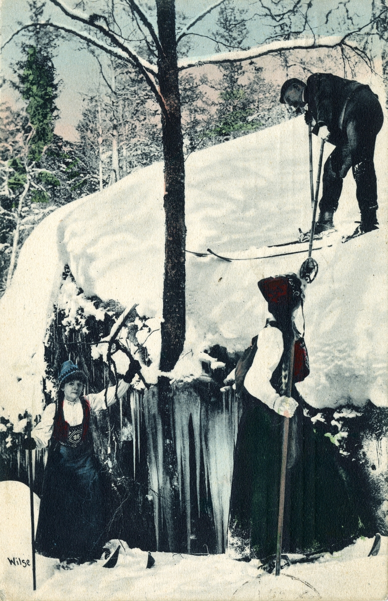 Kolorert postkort. En mann og to bunadskledde damer på ski skogen.