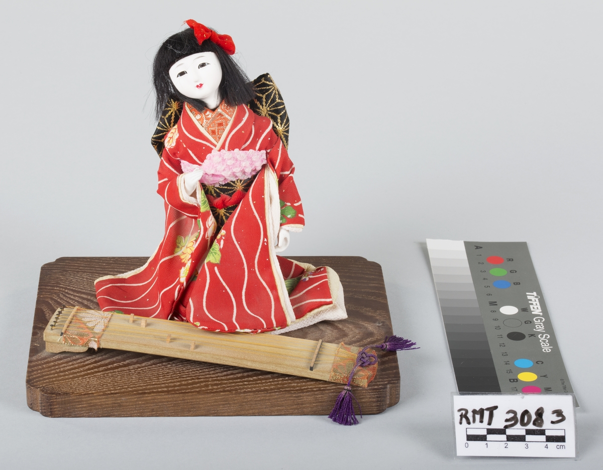 Sittende japansk dukke festet på et lavt platå av tre. Et rektangulært strengeinstrument, koto, foran seg.