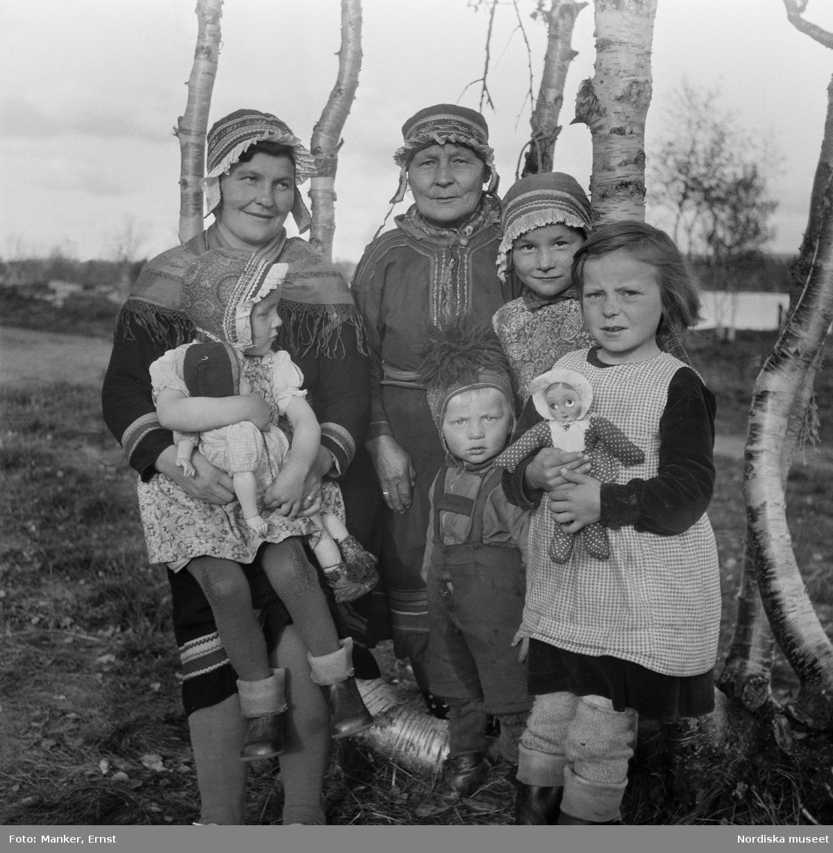 Porträtt av tre generationer samiska kvinnor och barn. Flickorna med dockor i famnen. Britta Prost (född Sevä 1912) med dottern Elin Ragnhild (född 1944), svärmor/faster Susanna Prost (född Sevä) och brorsbarnen Ellen Margareta och Karl Erling. Från Nordiska museets Lappmarksundersökning.