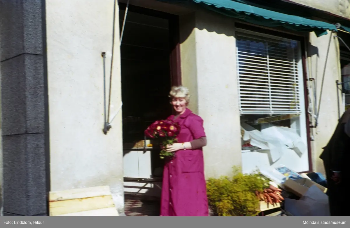 En kvinna står i dörren till något som skulle kunna vara en grönsaksaffär. Möjligen är adressen Kvarnbygatan 43 vid Gamla Torget i Mölndal, 1960-1970-tal.

För mer information om bilden se under tilläggsinformation.