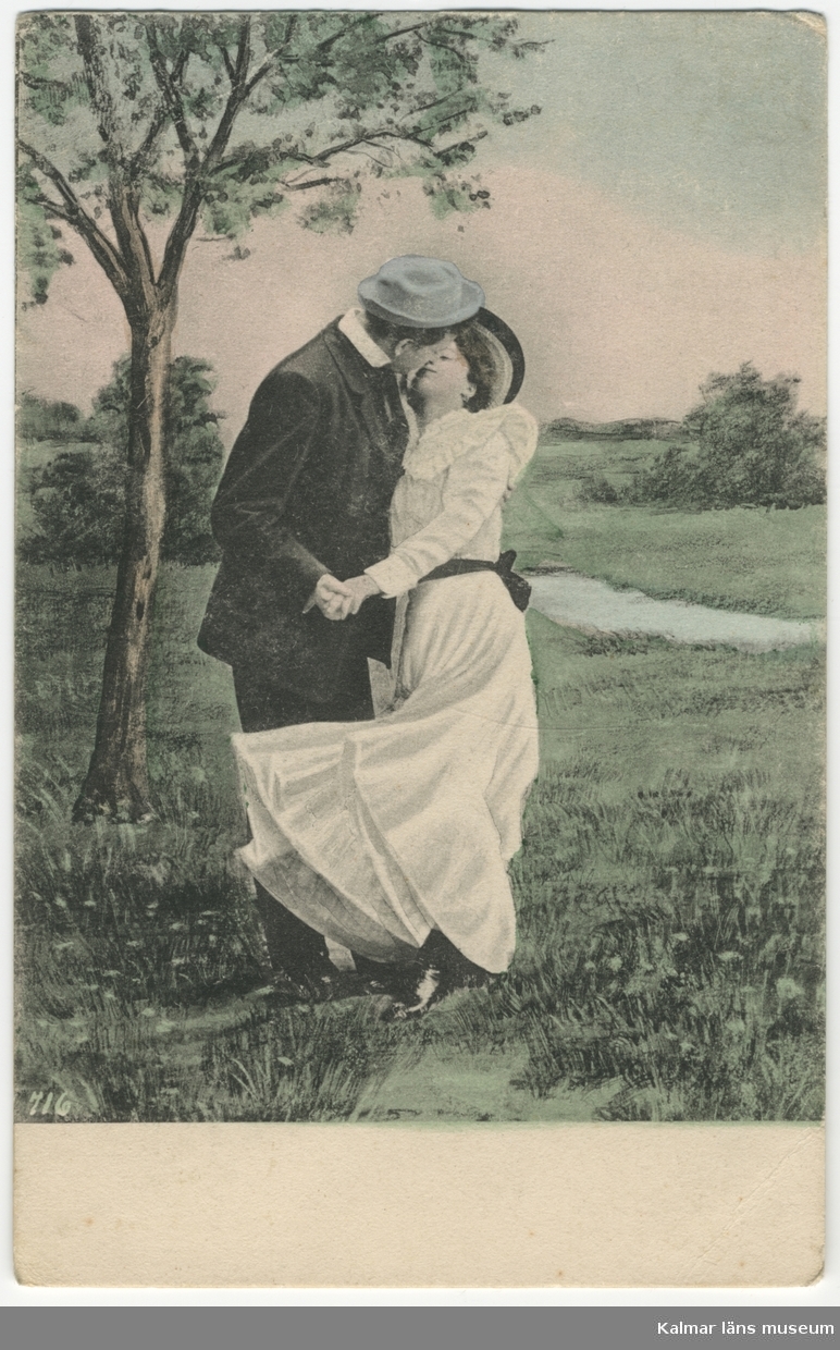 En man kysser en kvinna ute i en park eller liknande. Hon har hellång vit klänning.