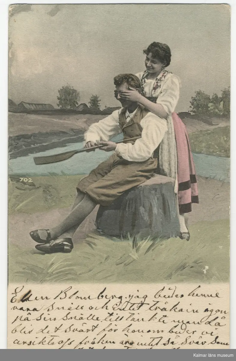 En ung man sitter på en stubbe och täljer på en åra eller skovel. En ung dam står bakom honom och håller för hans ögon. Jordbrukslandskap i bakgrunden.