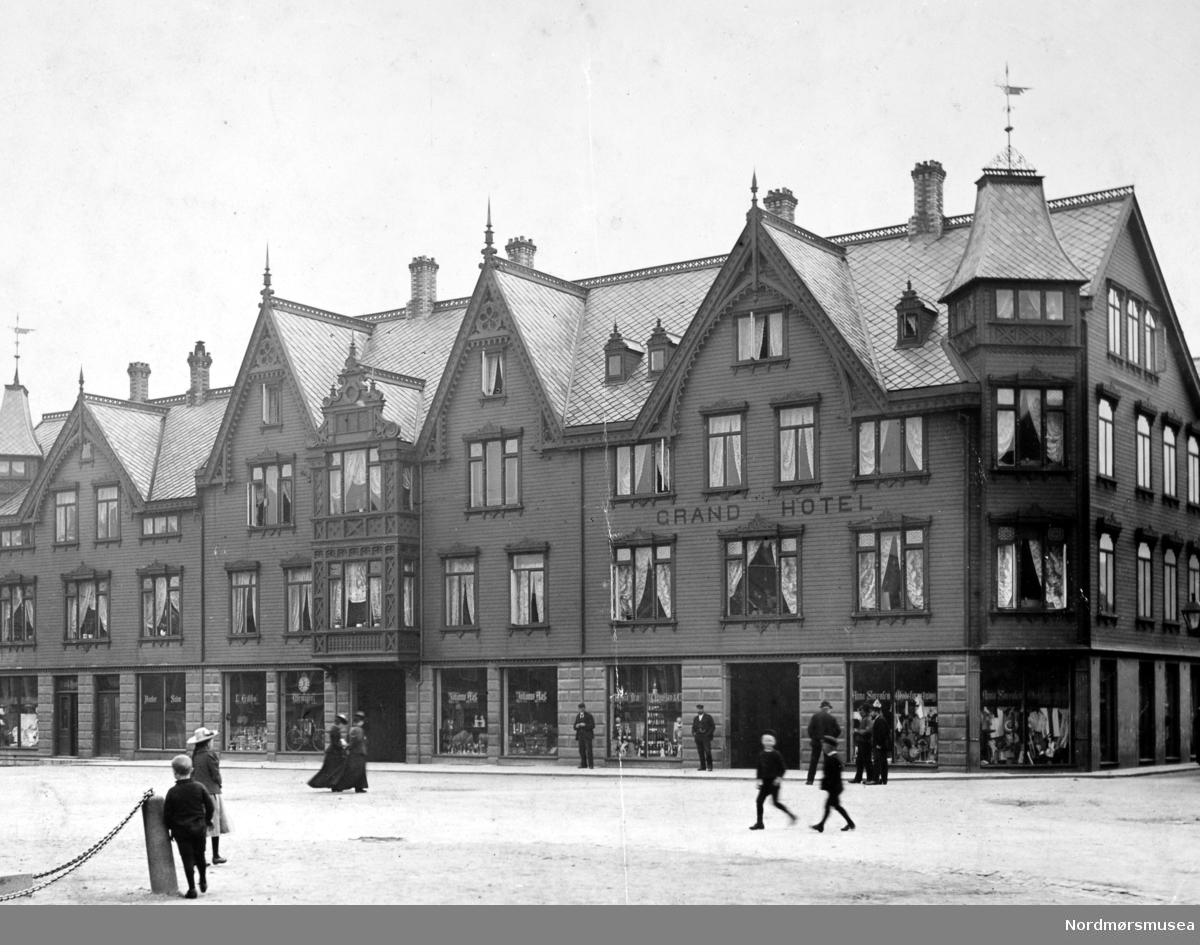 Det første Grand Hotell på Torget på Kirkelandet i Kristiansund.

Historie: Etter en alvorlig økonomisk krise i 1880 årene var byen nå atter preget av fremgang og optimisme. Byen hadde på denne tid en befolkning på 11 til 12000 innbyggere. Det var nå behov for et nytt standmessig hotell for reiselivsnæringen, foretningsfolk samt byens egen befolkning.

Gustav W. Dahl som var født i Trondheim i 1863, der han i første halvpart av 1890 årene hadde arbeidet som hotelltjener blant annet ved Grand Hotell cafe. At han ville prøve seg på hotelldrift i Kristiansund skyldes nok mye hans kone som var fra Kristiansund, og gjennom henne var blitt kjent med byen. 

Dette ledet til at i første halvpart av 1890-årene ble det oppført et boligkompleks på tomten til den gamle Prestegården, og som hadde fasade mot Torvet og Bernstorffstedet. Det var Stadskonduktør Hagbart Brinchmann som var byggherre og eier av bygget, hvor Grand Hotell etablerte seg i.

Mai 1895 kunne man lese i en annonse i to av byens aviser at byens Grand Hotell åpner dørene 1. juni 1895, og annonsen var signert hotellets eier Gustav W. Dahl. 

Hotellet hadde 30 værelser, og foruten gjesterom hadde det udpakningsværelser, en elegant Salon, Læseværelse og Spisesal.

Driften av hotellet ser ut til å ha godt bra, for i 1897 etablerte Gustav W. Dahl seg som restauratør på Øvre Bakklandet i Trondheim. Dette kjøpet ble fulgt opp av et nytt kjøp våren 1900, da han også kjøpte Trondheims Grand Hotell.

Ettersom Gustav W. Dahl nå hadde satset så hardt i Trondheim ble det behov for en ny innehaver for Grand Hotell i Kristiansund. 20. november 1900 kunne man da også lese i avisen i Kristiansund at nye lederen for hotellet nå var Nils Nilsen.

Nils Nilsen var født i Sand i Ryfylke i 1869. Han hadde vært reisefører og tolk for den britiske godseier og vinkongen Fleetwood Sandeman og gjennom hans reiser gjennom flere europeiske land, hadde fått et inngående kjennskap til hotelldrift. På det tidspunkt han tok over driften av hotellet var han faktor ved Sandemans gård Gulla i Surnadal. Trolig fikk Nils Nilsen også økonomisk støtte fra sin arbeidsgiver til å kjøpe hotellet.

En av forandringene Nils Nilsen gjorde ved hotellet var å installere hustelefon på alle rom.

En av de større begivenhetene hotellet opplevde var å være vertskap for kongefamilien på dens kroningsferd den 18. juni 1906. Nils Nilsen sørget først for en bedre borgermiddag i gymnastikksalen og i andre rom ved Enggatens skole. Hans Majestet Kong Haakon VII overnattet senere på hotellet, mens Hennes Majestet Dronning Maud overnattet på  kongeskipet Heimdal for å få mer ro neste morgen. Morgenen etter ble det arrangert en avskjedsfrokost for kongen, der medlemmer av formannskapet og embetsmenn med fruer deltok. 

Så i 1899 døde Hagbart Brinchmann som eide bygningen der Grand Hotell holdt til, og hans enke, Laura Susanne Brinchmann (født Øwre) solgte da bygningen til Nils Nilsen våren 1907. Nils Nilsen var nå blitt eier av både hotellet og bygningen.

Men så den 8. november 1907 brøt det ut brann i hotellets vedbu. Brannen ble oppdaget av nattevakten, og alarm ble slått. Mer enn 300 mann kom raskt til stede, samt 5 fra sjødampsprøyta, men det ble snart klart at det store trekomplekset ikke kunne reddes. To mennesker mistet livet i brannen. Disse var søstrene Anna og Augusta Sørensen som drev motehandel i første etasje av hotellet. Den flotte trebygningen i utført sveitserstil var nå blitt slukt av flammene.

Ikke lenge etter besluttet Nils Nilsen å bygge et nytt hotell på den samme tomten. Arkitekten ble Kristen Tobias Rivertz, og det nye hotellet som ble oppført i jugendstil, stod så ferdig januar 1909.

Referanse: En opplevelsens festlabyrint av Egil Husby, side 7 - 16. Nordmøre Museums fotosamling.
