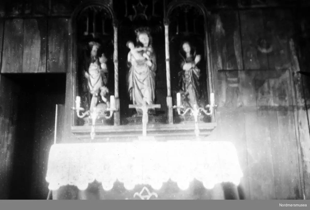 På bildet ser vi altret i Grip Kirke, med den hvite alterduken, lysestaker og krusifiks. Øverste del av av den sengotiske altertavlen med gotisk fletteverk og renessanse rundbuer er utydelige. En ser disse figurene fra venstre til høyre, utført i tre: 1.) Sankt Olav med rikseple i venstre og øksen i høyre hånd, som tråkker på en syndens figur, en drageaktig skikkelse, med menneskehode dekket av en turban (arketypen ";tyrkeren";). 2) Jomfru Maria med barnet på armen. 3.) St. Margaretha (på Grip kalt ";Maret";) - en ung kvinne med to lange fletter i buer. Et hunde- eller ulvehode stikker frem foran føttene hennes. Til venstre ser vi inngangen til sakristiet, til høyre skimter vi litt av veggmaleriet ";Abrahams offer";, med Abraham kledt i renessansedrakt, med engler over, samt med litt av Isaak til høyre for ham. BIldet er datert august(?) 1947. Litteratur: Tone M Olstad: Alterskapet i Grip stavkirke, NIKU Rapport 5, 2003. Jostein Iversen: Grip, 1998. Fra Nordmøre Museums fotosamlinger.
