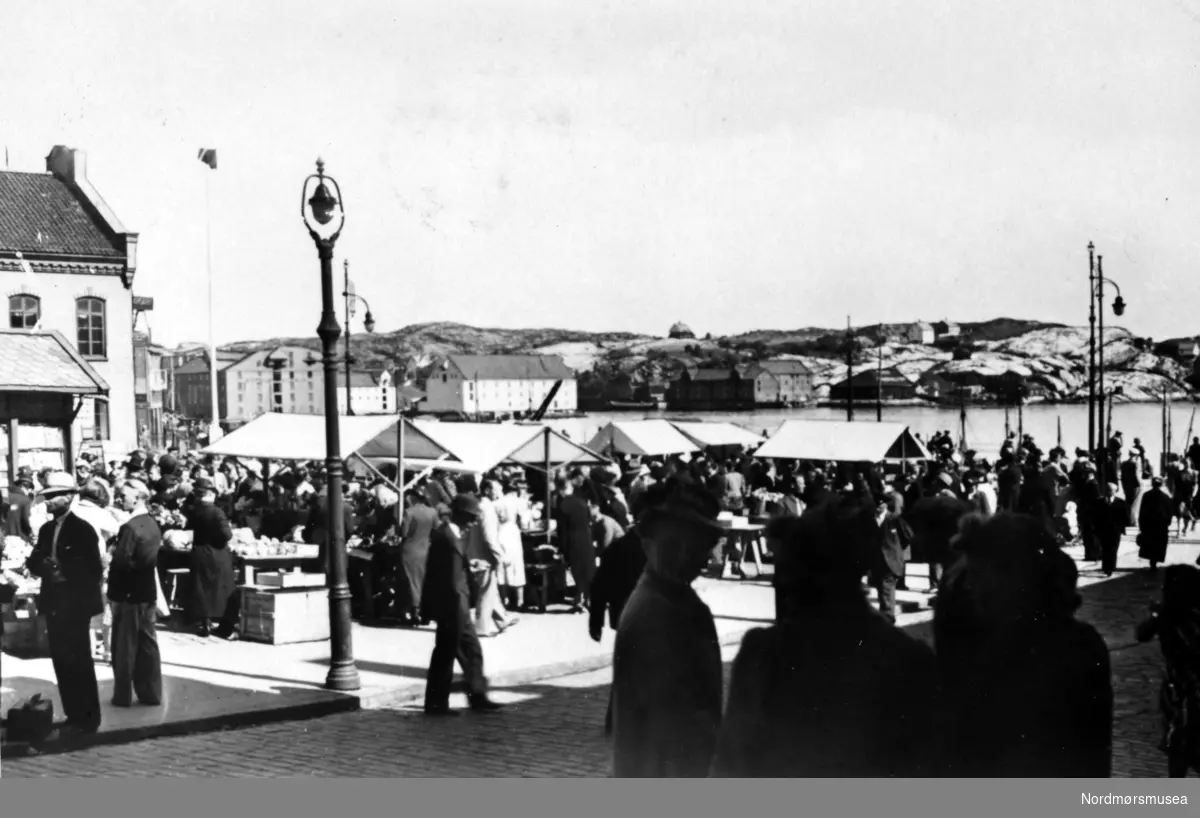 Livlig handel på Fisktorvet i Kristiansund i 1930-årene.  Midt i bildet ser vi havnebassenget. Til venstre i bildet ser vi litt av Tollboden og til høyre for flaggstangen ligger bryggene på Devoldholmen.  Til høyre i bakgrunnen ligger Gomalandet med Israelsneset og Milnbrygga med klippfiskbergene.
(Frå Nordmøre Museum si fotosamling)