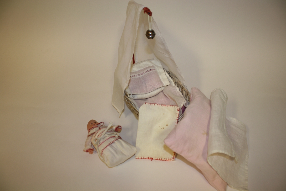 Celloloid-dukke i kurv med hjemmesydd sengetøy og sengeforheng og hjemmelaget stativ med bjelle. Dukken er reivet har har tøybleie og skjorte med krage.
