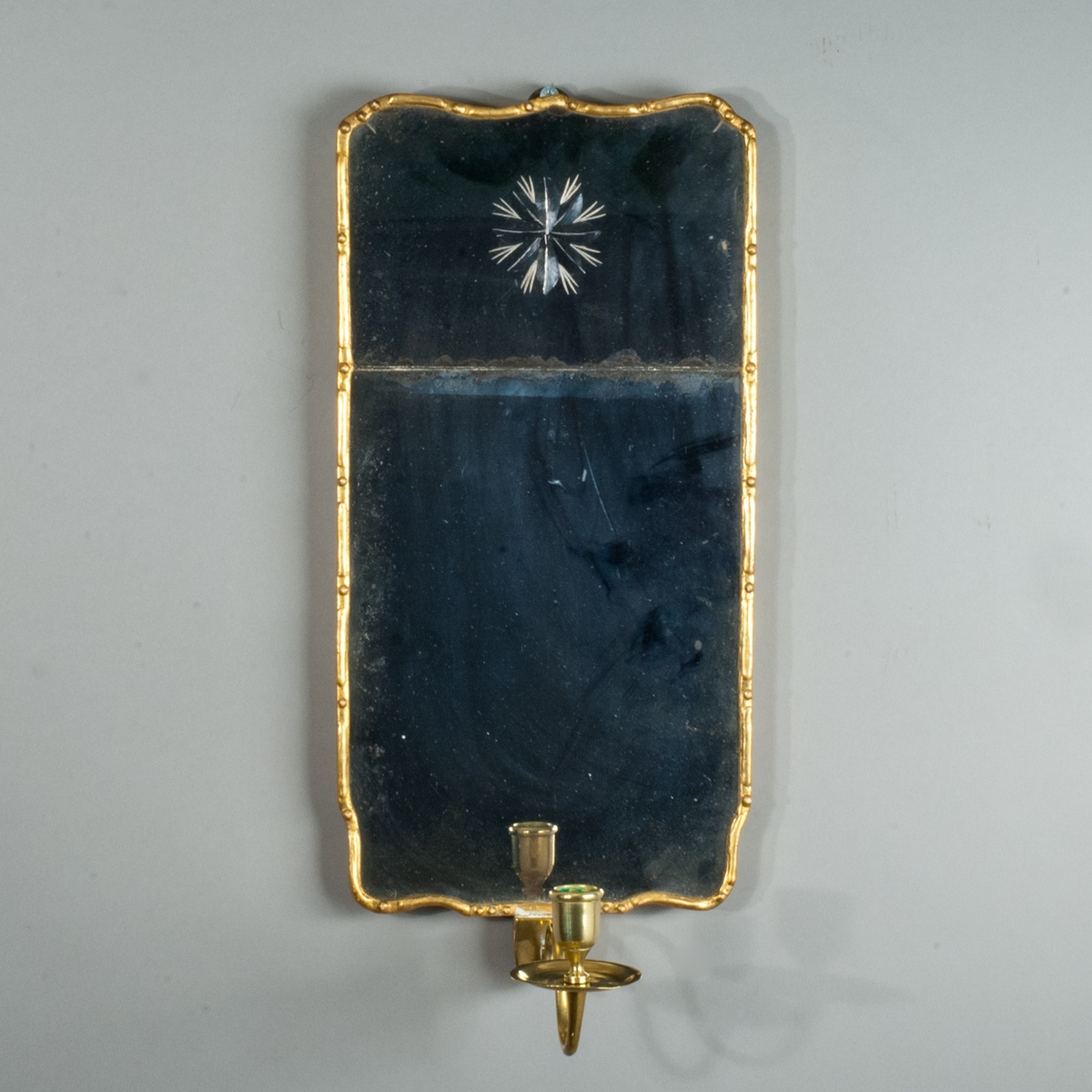 Lampett av spegelglas, rektangulär konturerad form med två glas infattade i tunn guldfärgad list med gles pärldekor. På det övre glaset en stjärna med strålar inslipad från baksidan. Bakstycke av trä. Ljusarm av mässing.