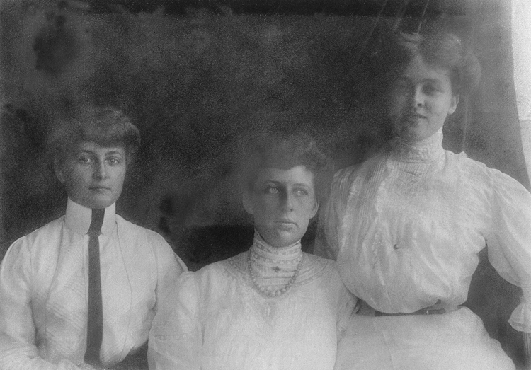 Närbild av tre kvinnor i vita, höghalsade sommarklänningar som sitter och står vid ett fönster.