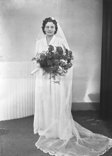 Foto av en brud. Hon är klädd i vit brudklänning, slöja och brudkrona. 
Helfigur. Ateljéfoto.