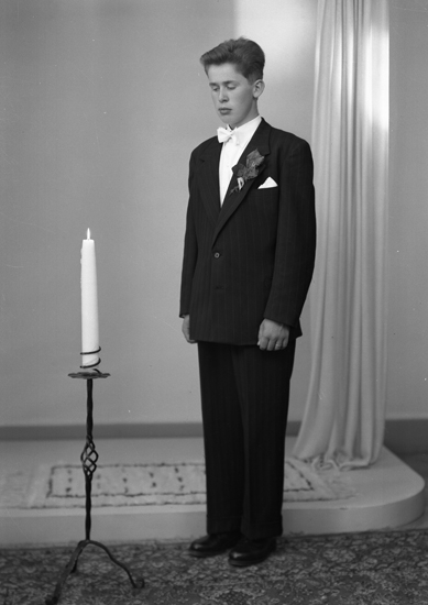 Foto av en ung man i mörk konfirmationskostym och vit fluga. Han står vid en tänd golvljusstake.
Helfigur, halvprofil. Ateljéfoto.
Konfirmand 1953. Lars Åke Snygg, Hjälmaryd, Blädinge, (1938 - ).
Son till Gunnar och Linneá Snygg.