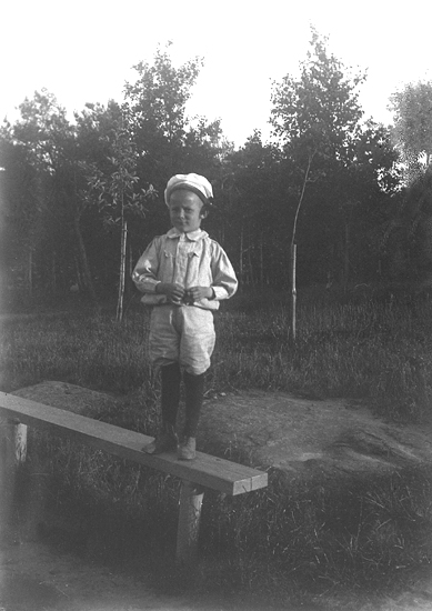 Foto av en pojke i knäbyxor m.m. På huvudet har han en rund, vit mössa. Han står på en trädgårdsbänk.