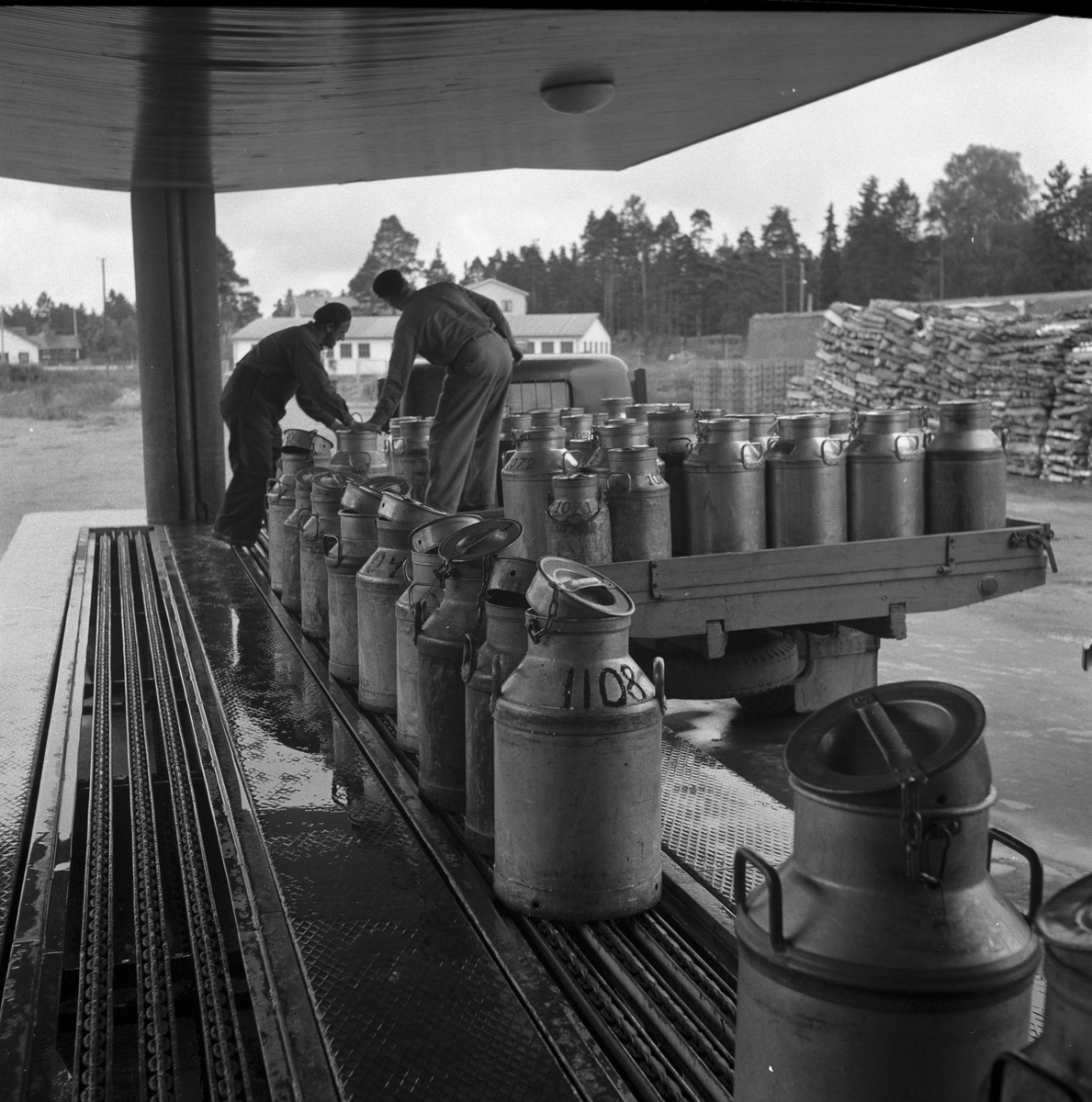Mjölkbilen från Järbo har anlänt till mejeriet. Herr Sigvard Andersson och Olle Jäderberg, Järbo, lastar av mjölkflaskorna på kajen vid Gävle Mejeri. De placeras på ett löpande band som går in i mejeriet.
Gävle Mejeri. 9 augusti 1953.
Reportage Gefle Dagblad.
