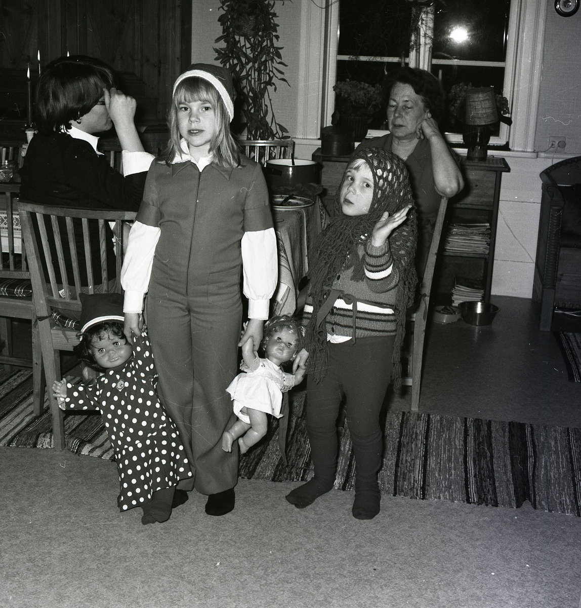 Kvinnor från flera generationers är samlade i ett rum. De äldre sitter vid bordet och samtalar med de yngre leker med två dockor i förgrunden. Flickorna är utklädda med luva och huvudduk när det fotograferas 1978 av Hilding Mickelsson.