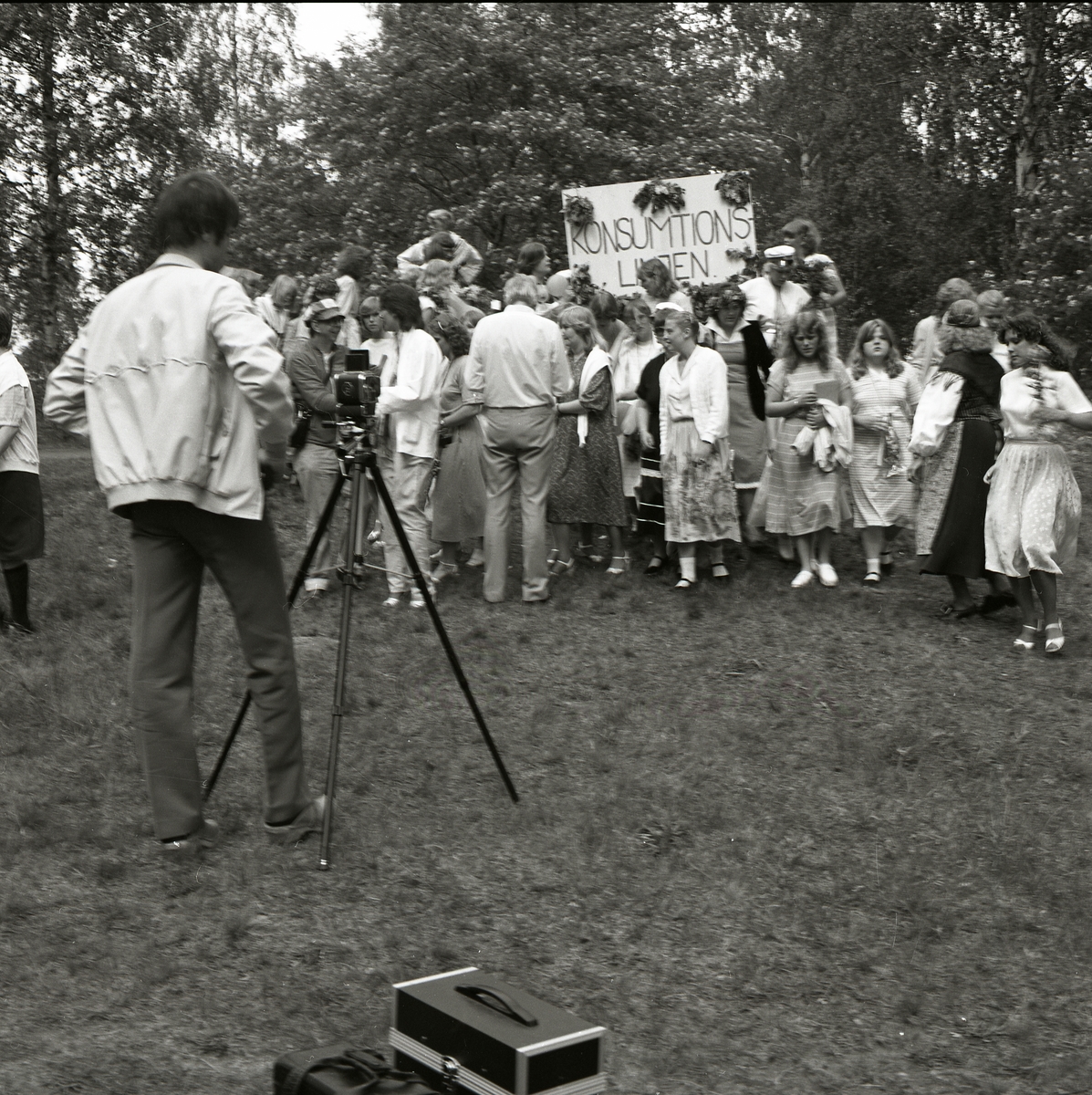 Konsumtionslinjen står skrivet på ett plakat dekorerat med blommor. Framför plakatet står en grupp med unga män och kvinnor i fina kläder och studentmössor. En fotograf står i förgrunden vid sitt stativ och har precis fotat gruppen som börjat skingra sig åt olika håll. Lövträden i bakgrunden bär sin fulla sommarskrud och skänker en sommarkänsla till bilden. Examensbilden är tagen i Bollnäs den 4 juni 1981.