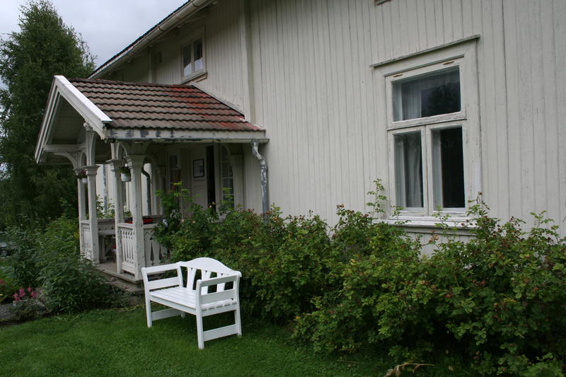 Det idylliske inngangspartiet til Sagstua skolemuseum og Sigurd Hoels barndomshjem. Hvitt bygg. Grønne busker langs husveggen.