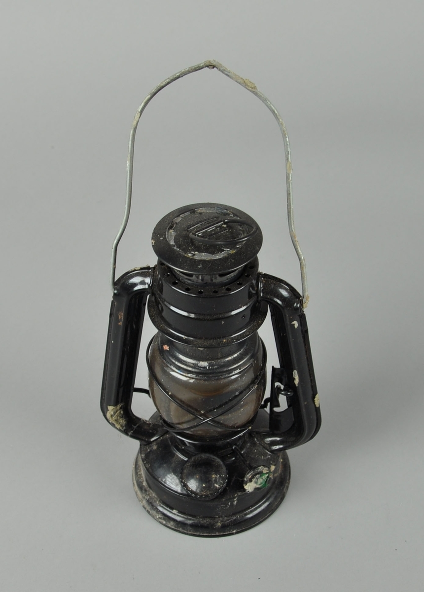 Oljelampe av metall med svart lakkering. Lampen har justerbart håndtak øverst, samt en hempe for oppheng.
