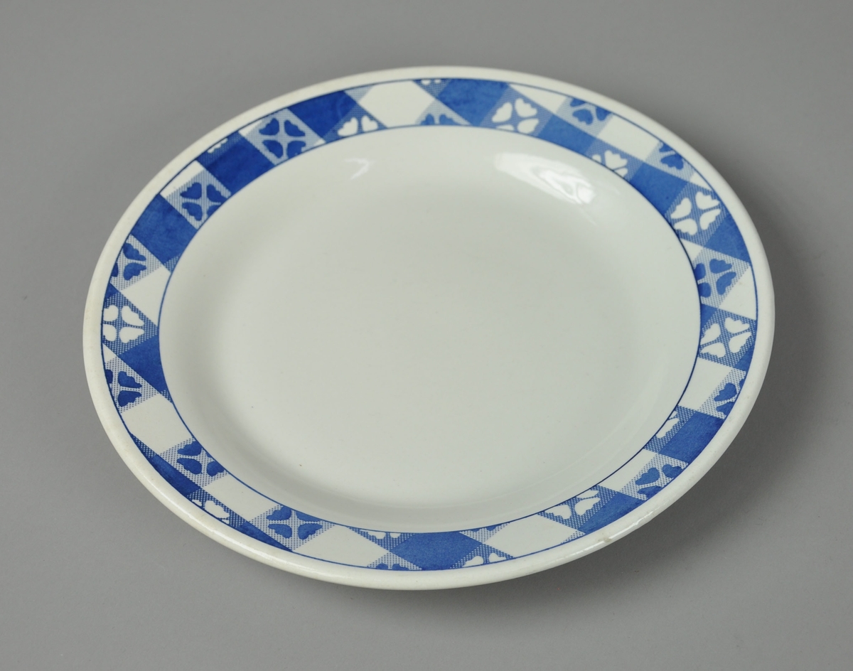 Tallerken av glassert keramikk. Rundt kanten går det en tykk borde med rutemønster inni og firkløvermotiv i rutene.