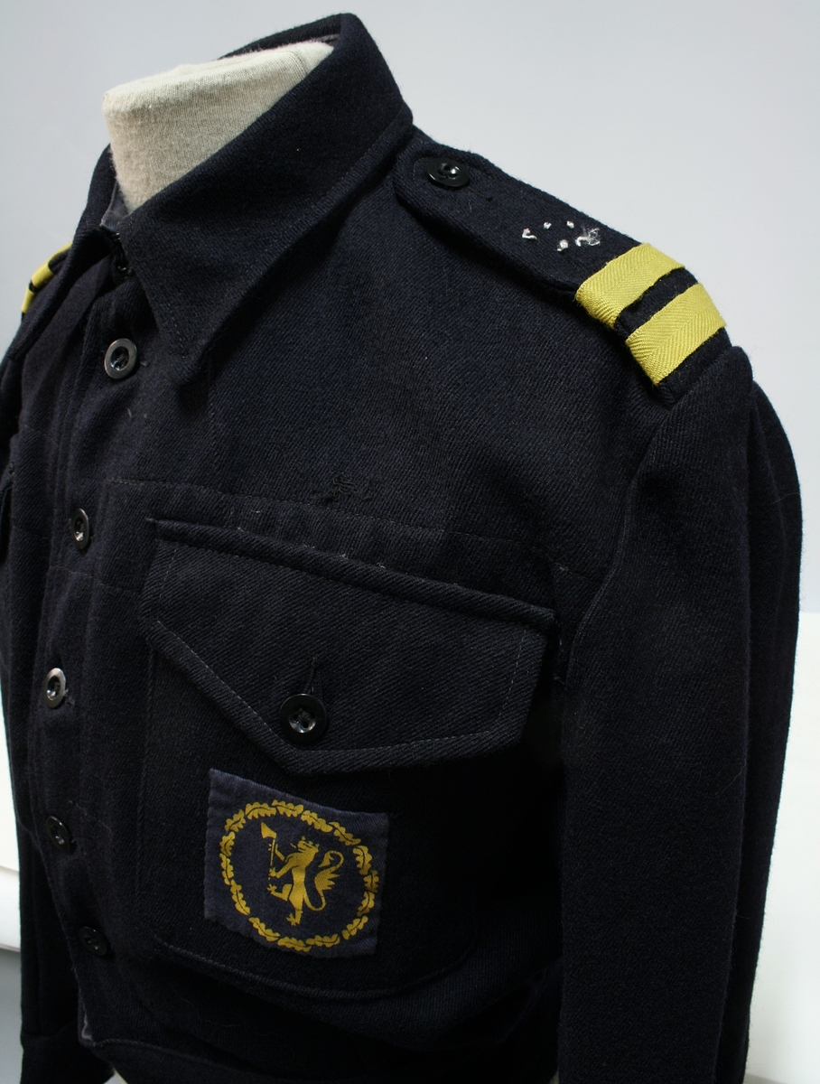 Mørkeblå battledress av den type som benyttes av de britiske A.R.P. Services. På venstre brystlomme et merke som viser den norske løve i en sirkelrund ekekrans, utført i gult på blå bunn.