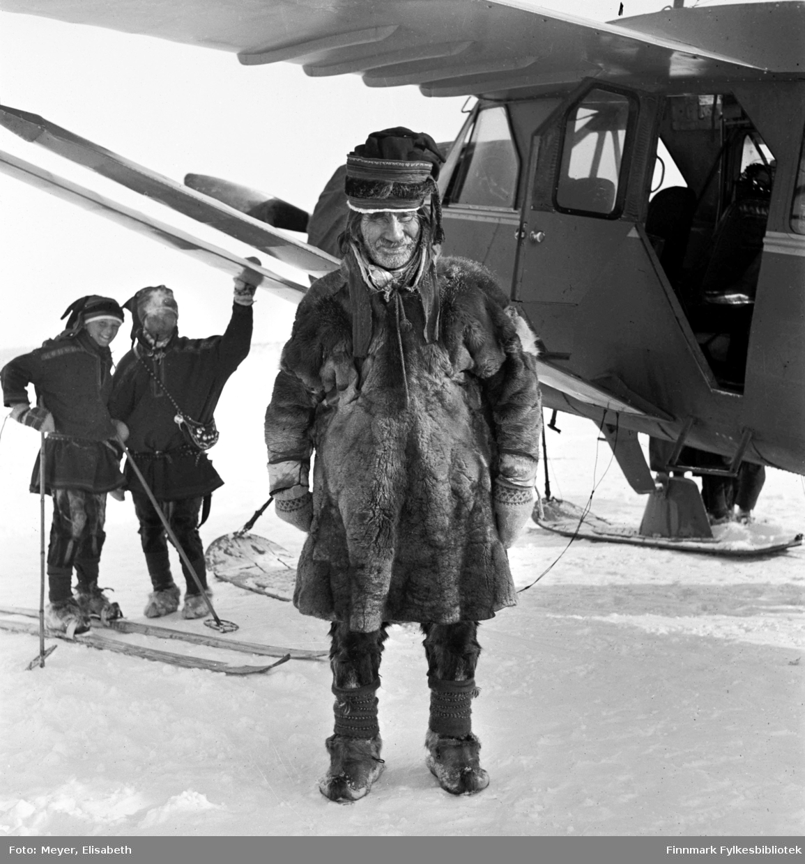 Johan Johansen Hætta foran et fly som har landet på Kautokeinoelva, fotografert i peridoen 1939-40 i Kautokeino. Johan Mathisen Hætta med pipe og gutt på ski står i bakgrunnen under vingen på  flyet.
