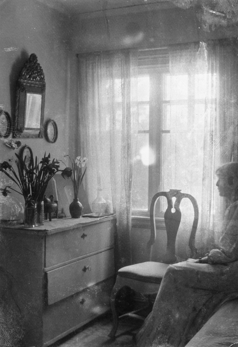 Ester Bauer sitter på sängen i blå rummet i stugan de hyrde i Torstorp utanför Gränna. Vaser med påsk- och pingsliljor står på en byrå mittemot henne.