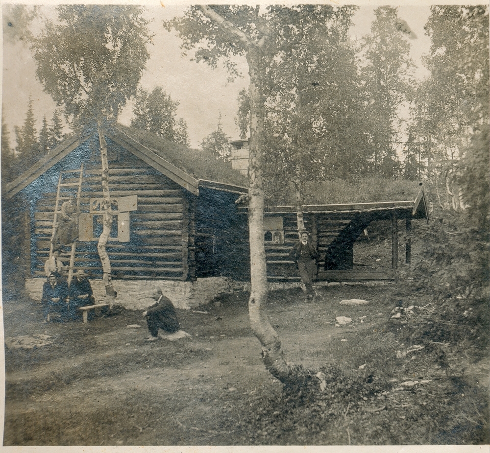 Gruppe. Utendørs. Personer ved tømmerhytte. En kvinne sitter i en stige.
Bildetekst: Baasmohytta 1910.
Oppl. tekst: Båsmohytta over Båsmosiden.