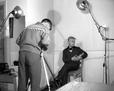 Fotostudio fra 1960-tallet. Ung mann med strikkegenser tar bilde av en annen ung mann i mørke klær.