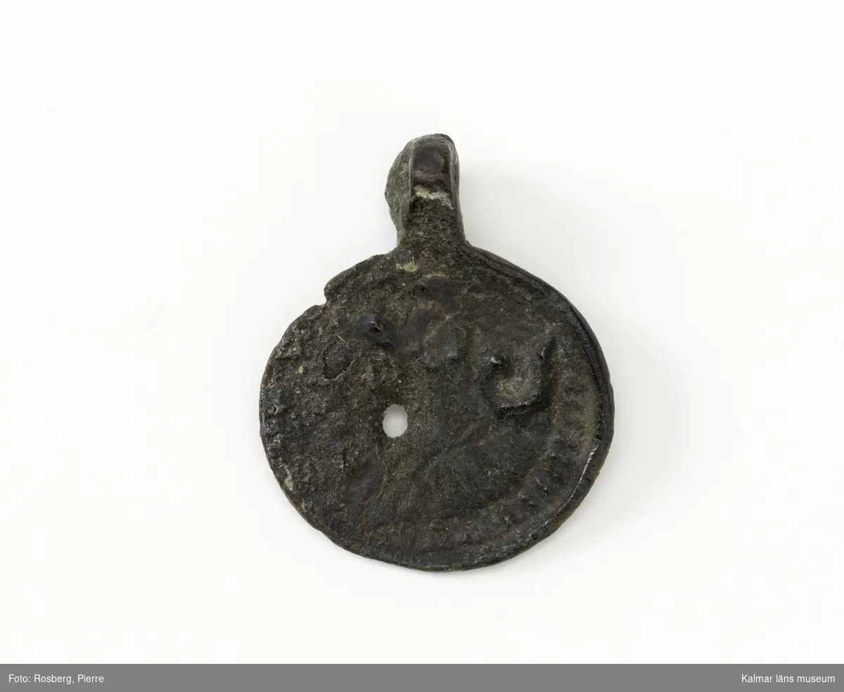 KLM 17748. Amulett?, hängsmycke, av metall. Tunn, rund platta med ögla. I plattan relief. Figur?.