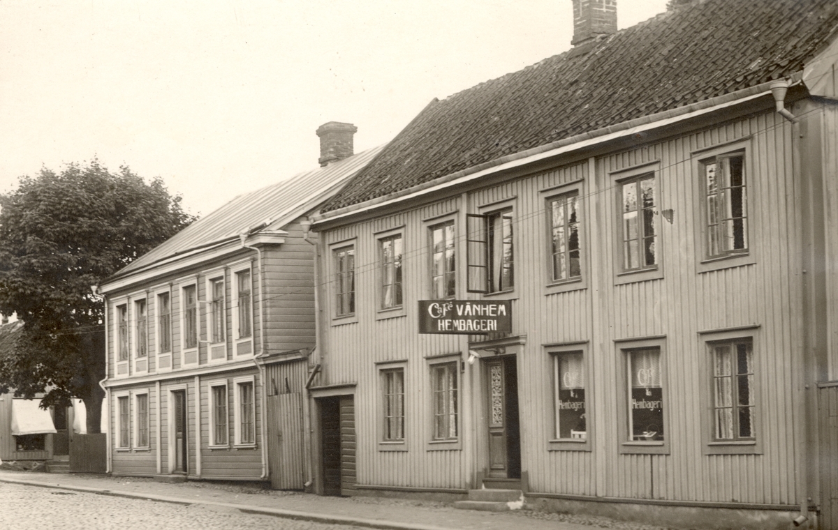 Två träbyggnader längs en gata, där det ena inrymmer ett kafé, Café Vänhem hembageri. 
På vykortets baksida finns en notering om att det är Hunnebergsgatan.