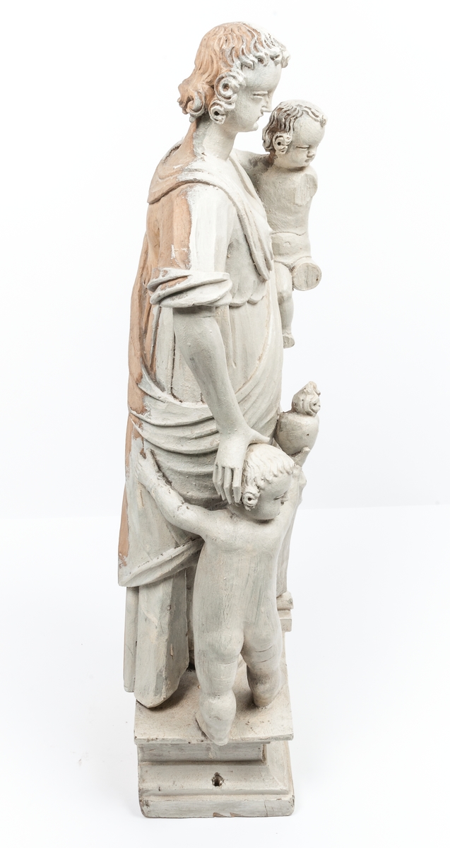 Kyrklig skulptur, trä, staty från gamla korskranket i Heliga Trefaldighets kyrka i Gävle, av Ewert Friis omkring 1665. 

Stående kvinnofigur med naket barn på vänster arm samt stående naket barn vid höger sida. Sistnämnda barn har ett brinnande hjärta i höger hand och kvinnans vänstra hand på sitt huvud. Vänster ben är främst, foten avslagen. Sittande barnets ena ben och andra fot avslagna. 
Fyrsidigt, profilerat postament. 
Gråvit färg.
