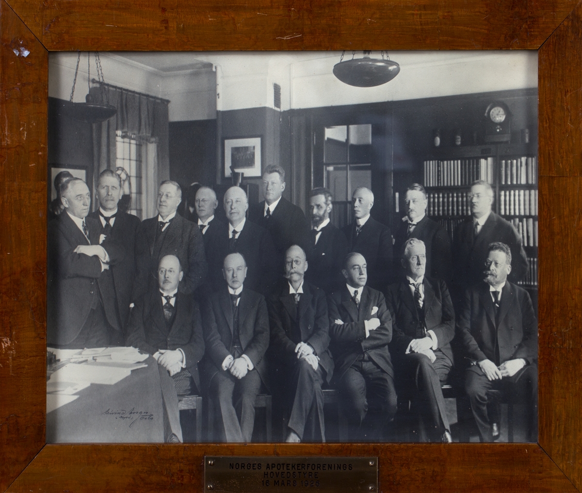 Gruppefoto av 16 voksne menn i dress. Seks sittende foran og 10 stående bak. En bokhylle bak til høyre i bildet.