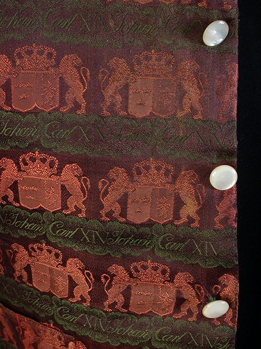 Med riksvapen och Carl XIV Johans namn i svart, rosa och grönt. Bakstycke av blå-brun-melerat bomullstyg. Foder av bomullslärft. Ståkrage.