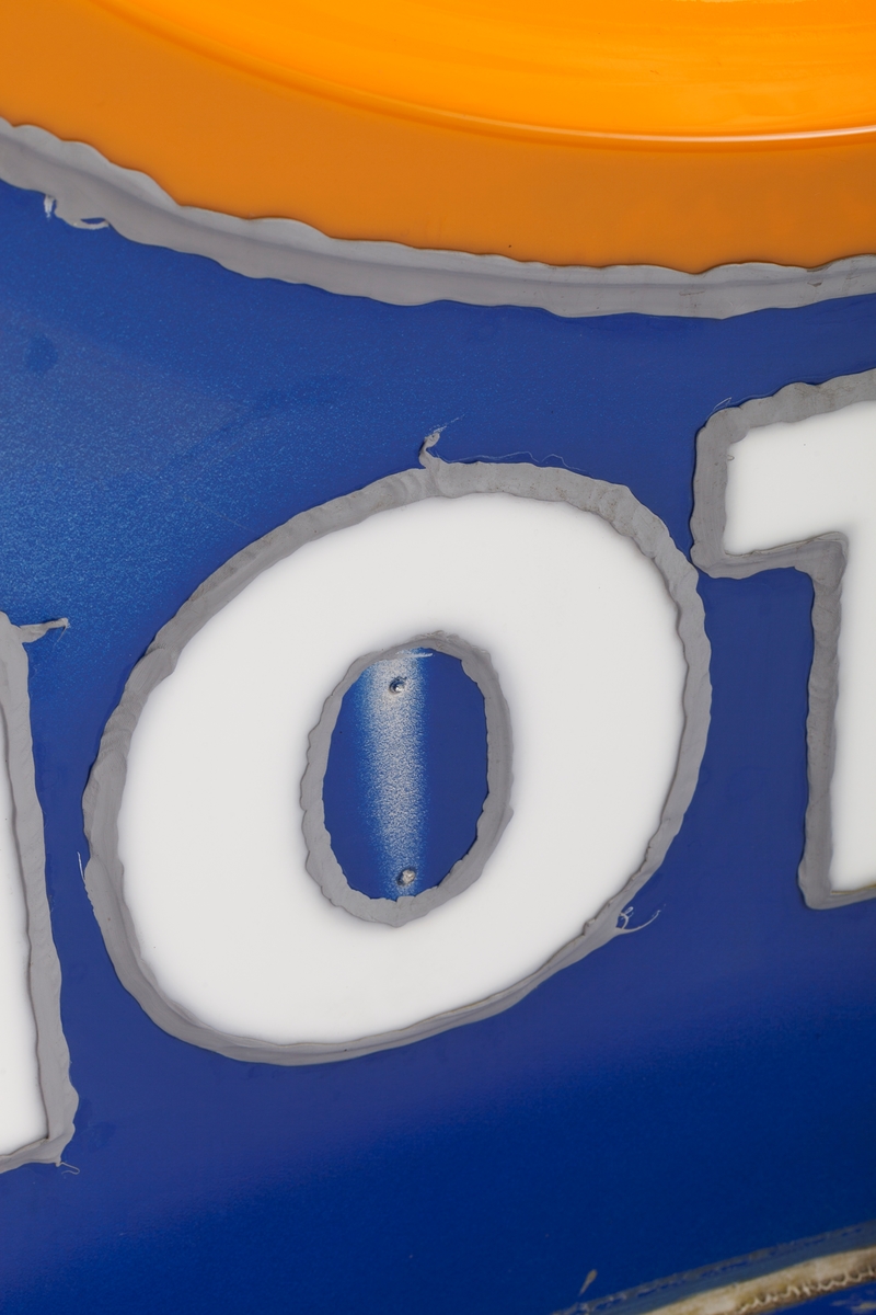 Skiltet med bakgrunnsfargen blå har en stilisert dråpe i oransje og det står Statoil med hvite bokstaver. Skiltet har vært del av et høyt veiskilt som har vært lyssatt, slik at det har lyst gjennom den oransje dråpen