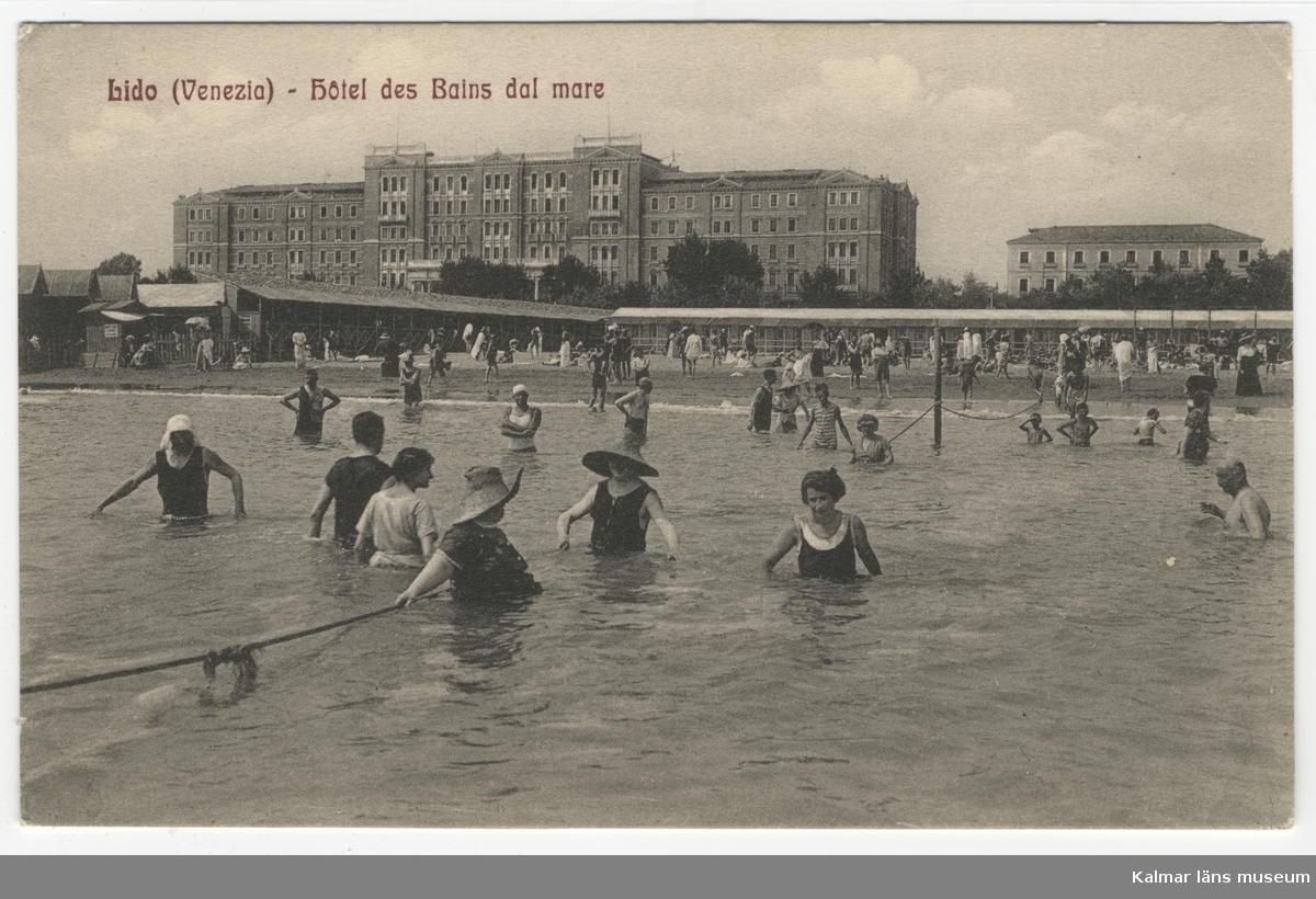 Badande män och kvinnor i mer eller mindre heltäckande badkläder, några bär bredbrättade hattar. I bakgrunden stranden och stora byggnader. Grand Hotel des Bains byggdes 1900.