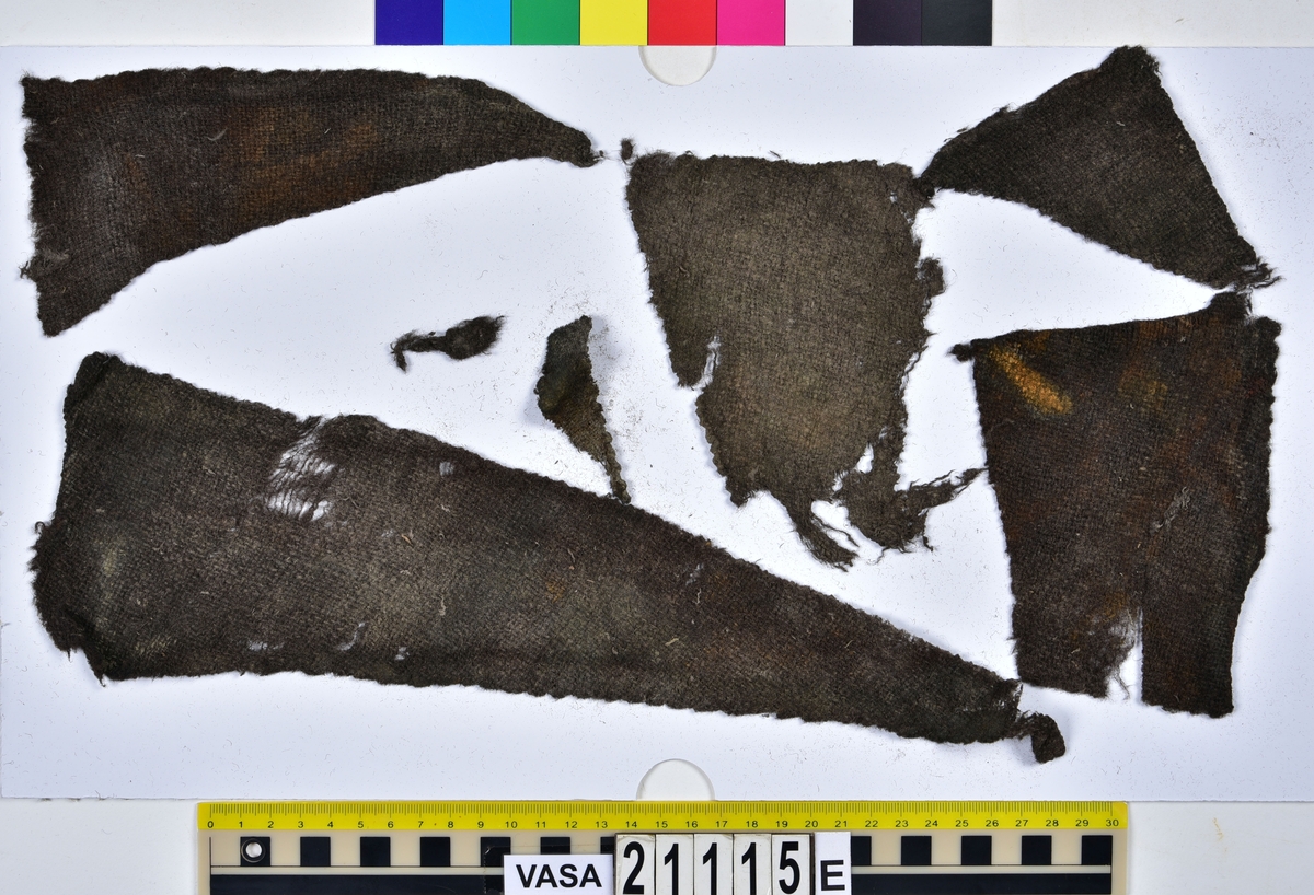 Fyndnummer 21115 består av en stor mängd olika textilier som hittades på undre batteridäck inuti tunna 21101 och var en del av den packning som fanns ombord. Vid utgrävningen av tunnan hittades textilfynden tillsammans med ett flertal andra personliga tillhörigheter så som skor, delar av en polsk stövel, nålbundna vantar av ull som hittades inuti vantar av läder, en hake, ett stort antal läderfragment, djurben, ett verktygsskaft och ett mynt.

Fyndnumret innehåller över 100 textilfragment av sex olika tyger fördelade i sju askar (a-g). Däribland finns delar till vad som kan ha varit ett ytterplagg så som en jacka, mandelka eller kasack sytt i ett ylletyg vävt i tuskaft. De bäst bevarade delarna av plagget består av två fram- eller bakstycken samt ett höger och ett vänster sidostycke. Tyget är något glest och har en del ojämnheter. Plagget är av en längre modell, där livdelarna och skörten är skurna i ett stycke utan söm i midjan. Fyndet har inga spår som tyder på att plagget har haft ärmar. På två fragment finns metallavtryck och spår av sömmar som visar att plagget har stängts med hjälp av hakar och hyskor.
Det finns även flera fragment från tre andra ylletyger vävda i kypert i fyndnumret. Dessa tyger är tätare och av en finare kvalitet än det tuskaftsvävda. Ett av dessa tyger ligger i form av mycket nedbrutna fragment på de större plaggdelarnas båda sidor. Utifrån tygernas olika kvaliteter så är det möjligt att det bättre bevarade grövre tyget utgjort plaggets foder medan tygen av finare kvalitet har varit plaggets yttertyg. Detta kan vara anledningen till varför tygerna har bevarats så olika då det kypertvävda tyget på så sätt har fungerat som ett slags skydd för det tuskaftsvävda tyget. Under 1600-talet användes ett glesvävt ylletyg kallat Boj som foder, vilket är en tygkvalitet som överensstämmer med fyndets glesare tuskaftsväv.

För mer detaljerad information från textildokumentationen (år 2022) se länkade filer.