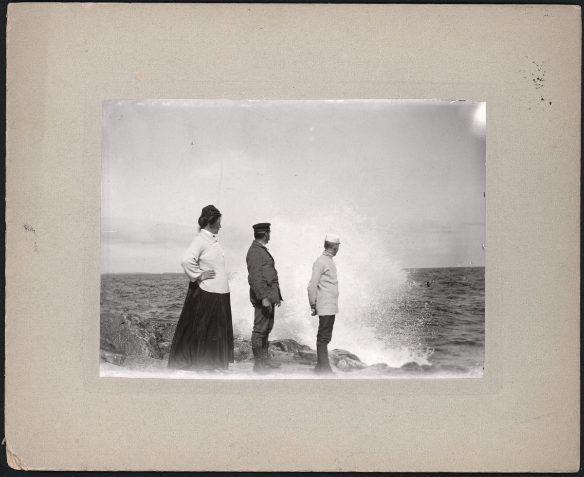 En kvinna och två män på en klippa vid havet, i bakgrunden slår en våg upp. "'Tripp, Trapp, Trull' försjunkna i beundran af hafsscenerier vid Landsort den 24 aug. 1905".