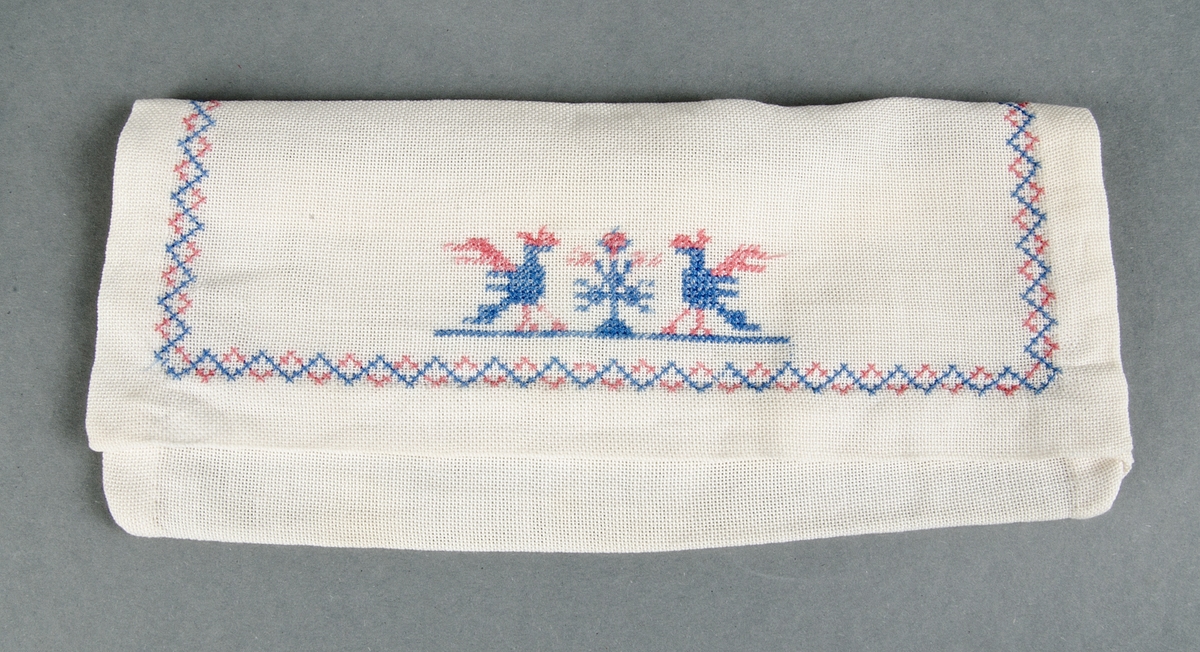 Servettväska av vitt glest tuskaftat bomullstyg handsydd med korsstygnsbroderi i merceriserad bomullstråd, två fåglar och bård i blått och rött.

