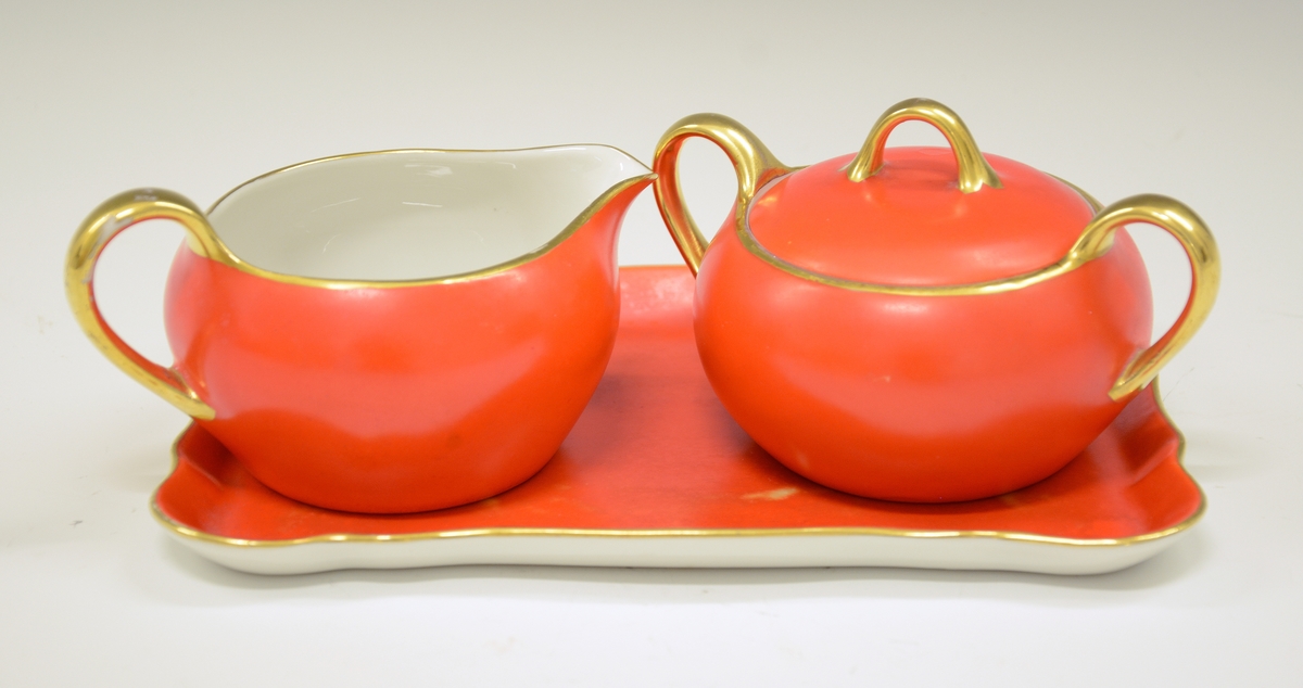 Sukkerkopp med lokk, av porselen. To hanker. Hvit glasur. Oransje med gullkant og gullhanker på kopp og lokk.
Modell 1490.